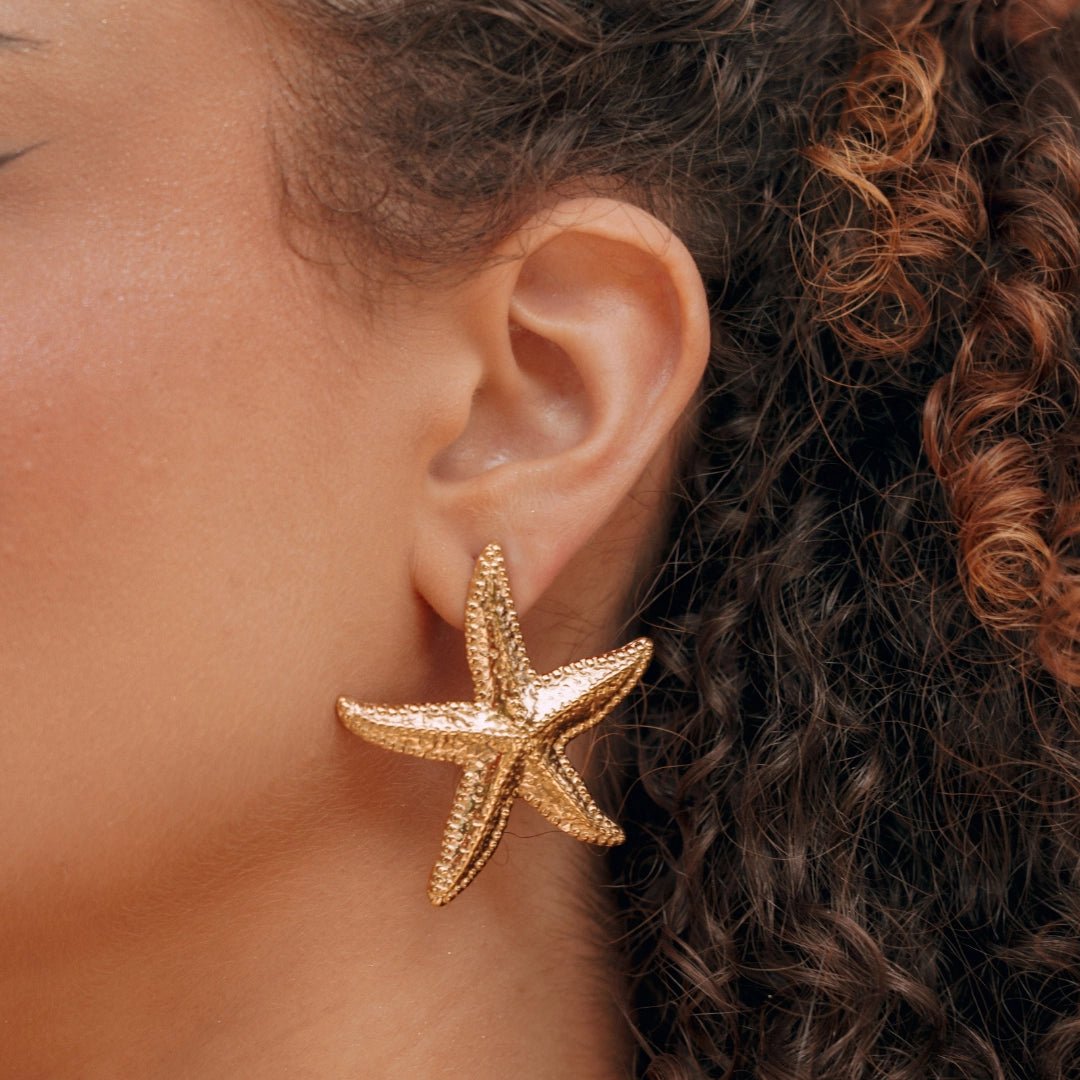 Boucles d'oreilles étoile de mer dorées en acier inoxydable avec texture détaillée, portées sur l'oreille d'une femme.