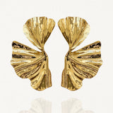 Paire de boucles d'oreilles larges motif abstrait feuille plissée dorée sur fond blanc.