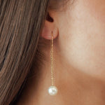 Boucles d'oreilles traversantes (ou enfile-aiguille) avec chaîne fine dorée et perle blanche pendante, style minimaliste et chic.