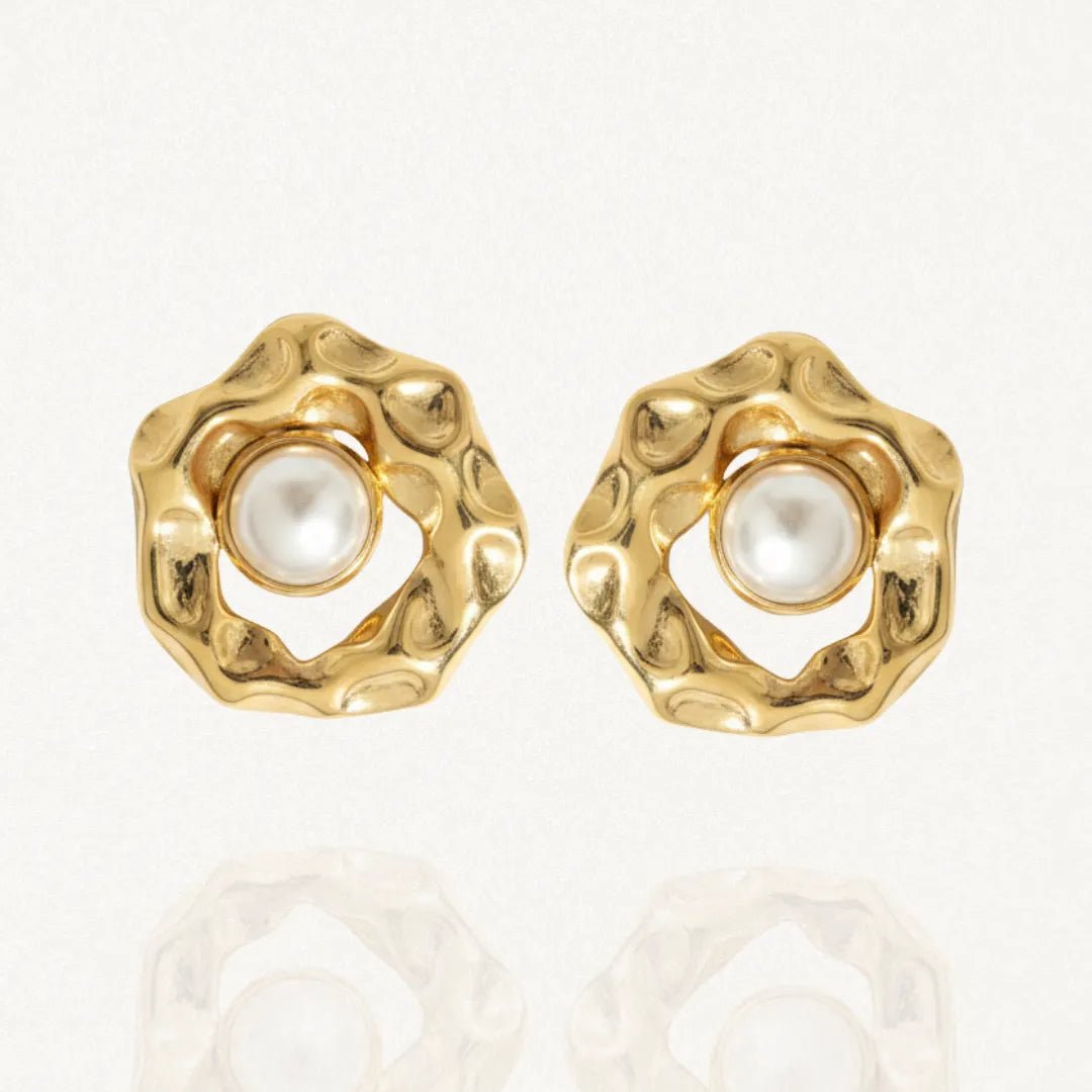 Paire de boucles d'oreilles dorées de forme organique texturée, avec une perle blanche au centre, posées sur un fond blanc réfléchissant.