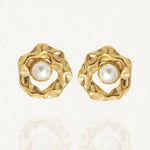 Paire de boucles d'oreilles dorées de forme organique texturée, avec une perle blanche au centre, posées sur un fond blanc réfléchissant.