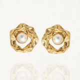 Paire de boucles d'oreilles dorées de forme organique texturée, avec une perle blanche au centre, posées sur un fond blanc réfléchissant.