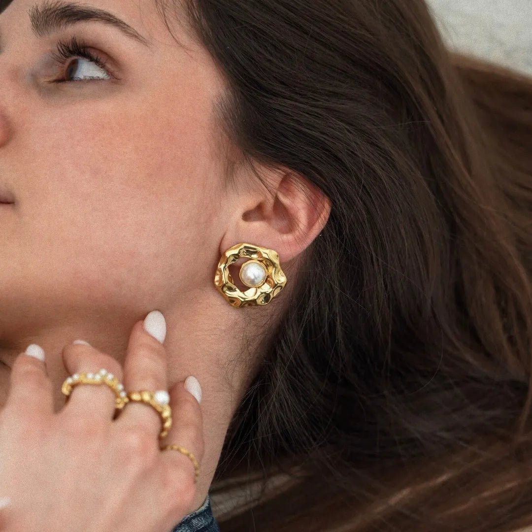 Boucle d'oreille dorée à texture organique et perle blanche portée par une femme avec bagues dorées et perles.