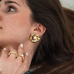 Boucle d'oreille dorée à texture organique et perle blanche portée par une femme avec bagues dorées et perles.