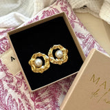 Paire de boucles d'oreilles dorées martelées ondulées avec perles blanches centrales, présentées dans un écrin sur tissu à motifs.