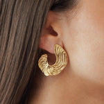 Boucles d'oreilles statement grand format forme vague ondulée et texturée en acier inoxydable doré portées à l'oreille.