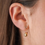 Gros plan sur la boucle d'oreille Harmonie : le clou cristal sur le lobe et la goutte dorée suspendue en dessous.