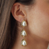 Boucles d'oreilles longues pendantes composées de trois gouttes blanches effet perle et contour doré portées à l'oreille.