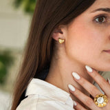 Femme portant une boucle d'oreille cœur dorée striée, une bague dorée large à bord ondulé avec nacre au doigt, chemise blanche.