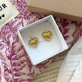 Deux boucles d'oreilles cœur dorées facettées présentées dans une boîte à bijoux blanche, sur tissu matelassé rose.