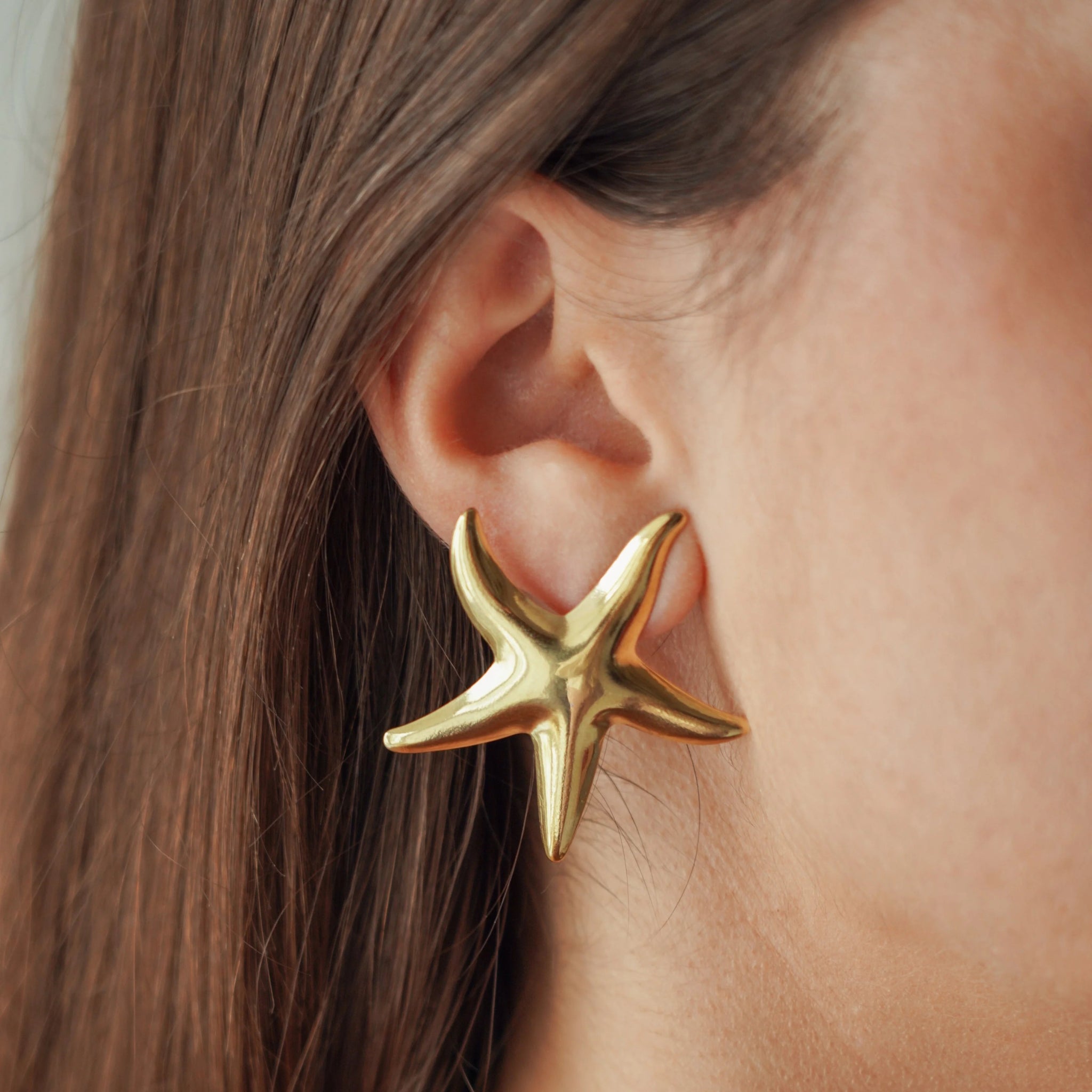 Boucles d'oreilles puces grand format motif étoile de mer en acier inoxydable doré portées à l'oreille, style vacances d'été.