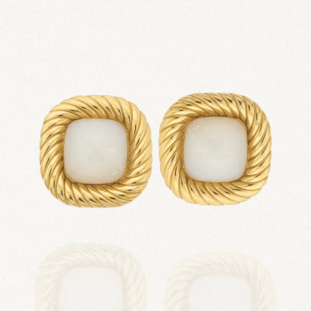 Une paire de boucles d'oreilles dorées carrées, avec un contour torsadé et un cabochon central en résine blanche opaque, posées sur un fond blanc.