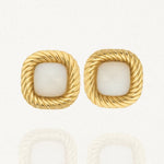 Une paire de boucles d'oreilles dorées carrées, avec un contour torsadé et un cabochon central en résine blanche opaque, posées sur un fond blanc.