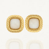 Une paire de boucles d'oreilles dorées carrées, avec un contour torsadé et un cabochon central en résine blanche opaque, posées sur un fond blanc.