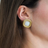 Boucle d'oreille carrée dorée à bord torsadé avec cabochon central blanc opaque, portée sur l'oreille d'une femme.