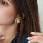 Gros plan sur une boucle d'oreille carrée dorée torsadée avec cabochon blanc laiteux, portée par une femme aux cheveux bruns.