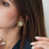 Gros plan sur une boucle d'oreille carrée dorée torsadée avec cabochon blanc laiteux, portée par une femme aux cheveux bruns.