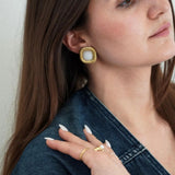 Boucle d'oreille carrée or à bord torsadé et pierre centrale blanche, portée par une femme en veste denim.