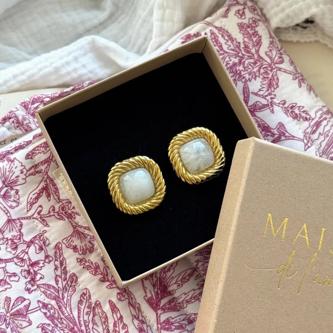 Deux boucles d'oreilles dorées carrées, cabochon blanc marbré, bord torsadé, dans une boîte beige avec insert noir, sur tissu floral.