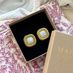 Deux boucles d'oreilles dorées carrées, cabochon blanc marbré, bord torsadé, dans une boîte beige avec insert noir, sur tissu floral.