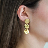 Oreille de femme portant une boucle d'oreille pendante dorée. Elle se compose de 3 disques, le supérieur lisse, les deux autres martelés.