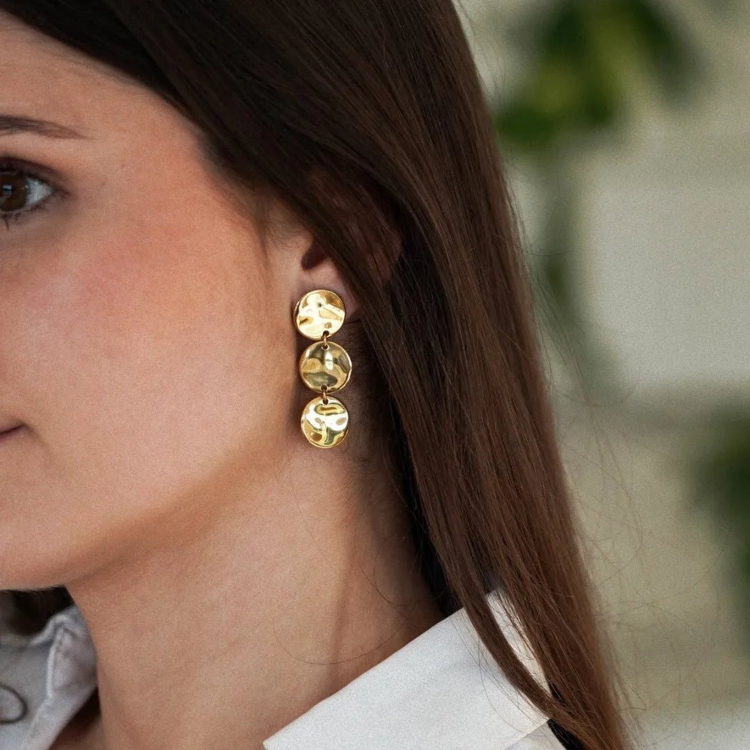 Boucle d'oreille dorée pendante à trois disques ronds martelés portée par une femme.