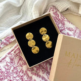 Boucles d'oreilles dorées pendantes avec 3 disques ronds martelés, présentées dans un écrin marron sur tissu blanc et fuchsia.