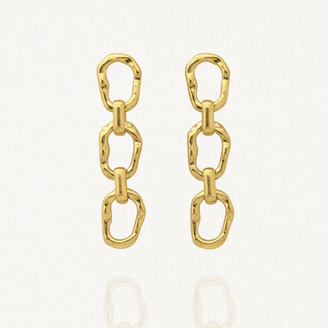 Paire de boucles d'oreilles pendantes dorées, composées de trois maillons ovales allongés à la surface irrégulière et texturée, sur fond blanc avec un léger reflet.