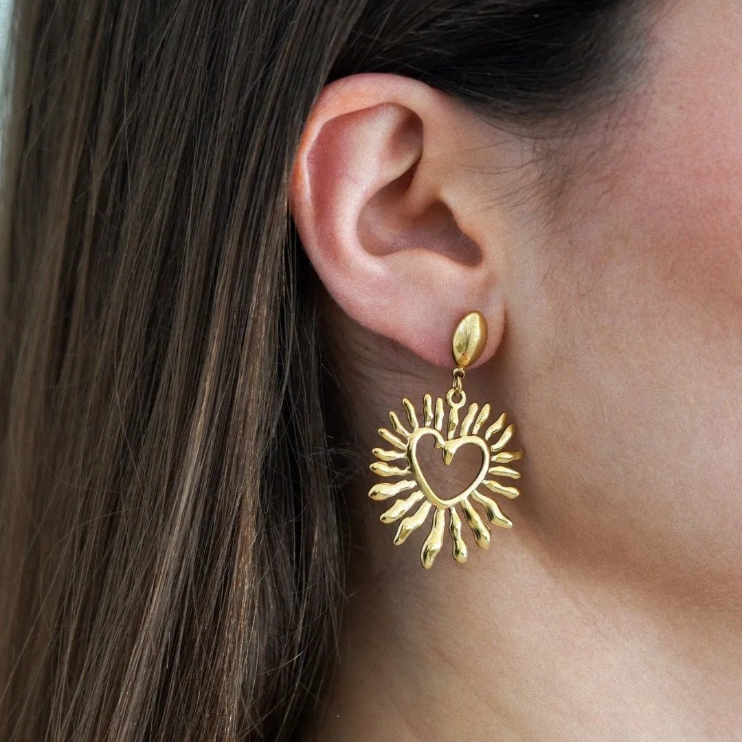 Gros plan oreille femme avec boucle d'oreille dorée pendante, motif soleil rayonnant avec cœur découpé.