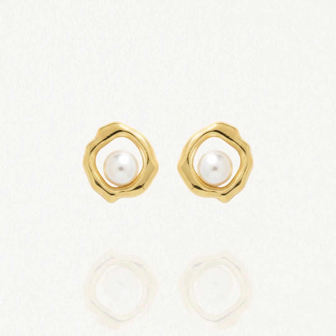Paire de boucles d'oreilles clous de couleur dorée avec un contour ondulé et une perle blanche ronde au centre de chaque boucle, posées sur un fond blanc réfléchissant.