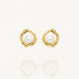 Paire de boucles d'oreilles clous de couleur dorée avec un contour ondulé et une perle blanche ronde au centre de chaque boucle, posées sur un fond blanc réfléchissant.