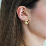 Boucle d'oreille dorée organique avec perle blanche centrale, portée sur l'oreille droite d'une femme aux cheveux bruns.