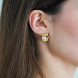 Boucle d'oreille dorée organique avec perle blanche centrale, portée sur l'oreille droite d'une femme aux cheveux bruns.