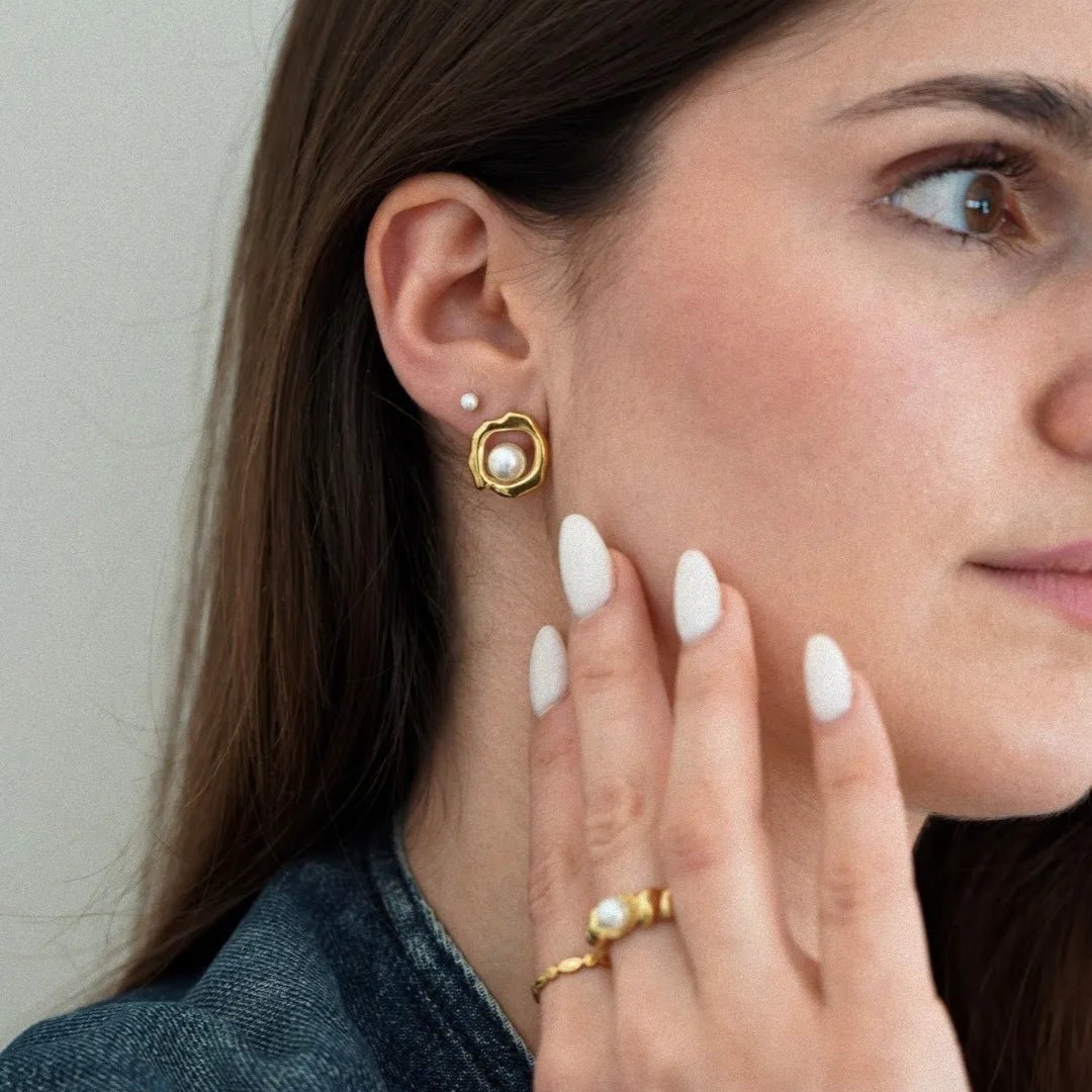 Clou d'oreille or forme organique avec perle, petit clou perle, et bague or avec perle, portés par femme aux ongles blancs.