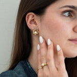 Clou d'oreille or forme organique avec perle, petit clou perle, et bague or avec perle, portés par femme aux ongles blancs.