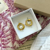 Boucles d'oreilles dorées, design organique irrégulier, avec perle blanche au centre, dans une boîte bijou blanche.