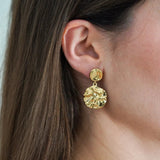 Boucle d'oreille pendante dorée martelée à formes irrégulières sur l'oreille d'une femme aux cheveux châtains.