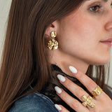 Femme avec boucle d'oreille dorée martelée, bague croix pavée de pierres et bague large en or texturé.