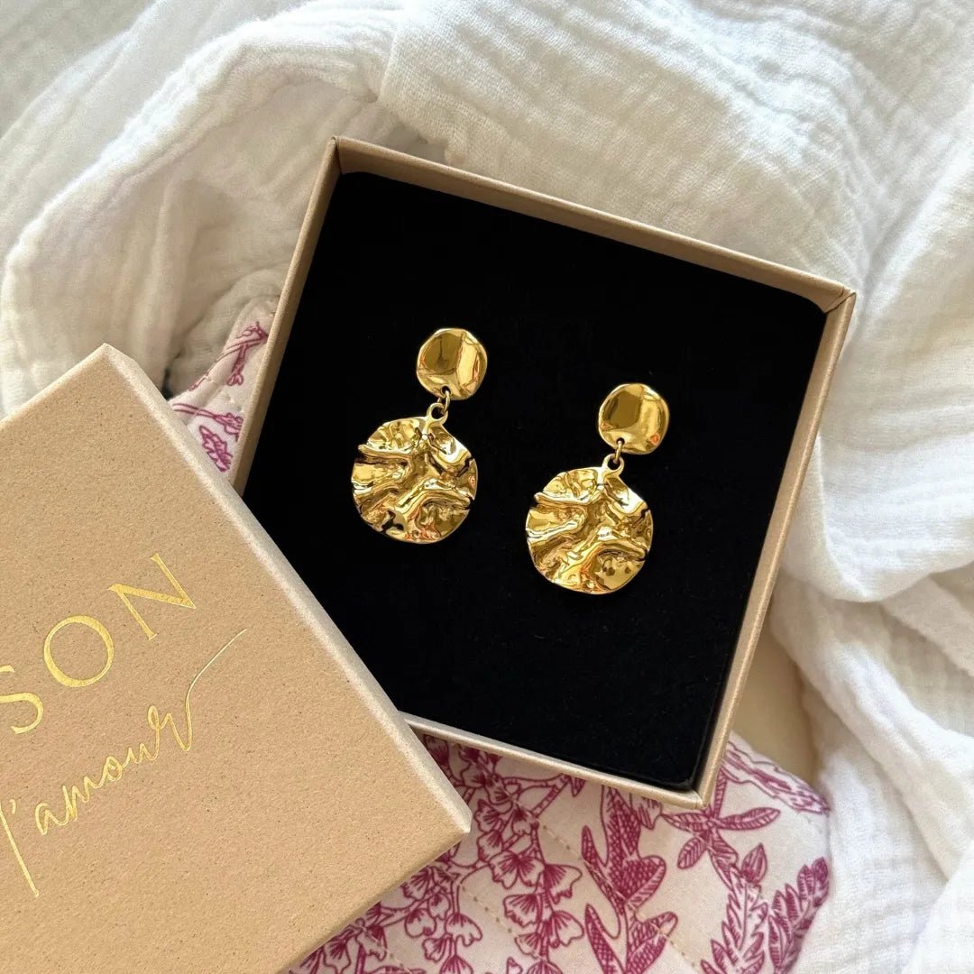 Boucles d'oreilles pendantes dorées avec stud ovale lisse et disque froissé texturé, dans une boîte cadeau beige ouverte.