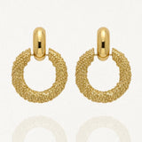 Paire de boucles d'oreilles pendantes dorées, composées d'une créole épaisse avec une texture granulée, suspendue à une attache ovale lisse et brillante. L'image est sur fond blanc avec des reflets.