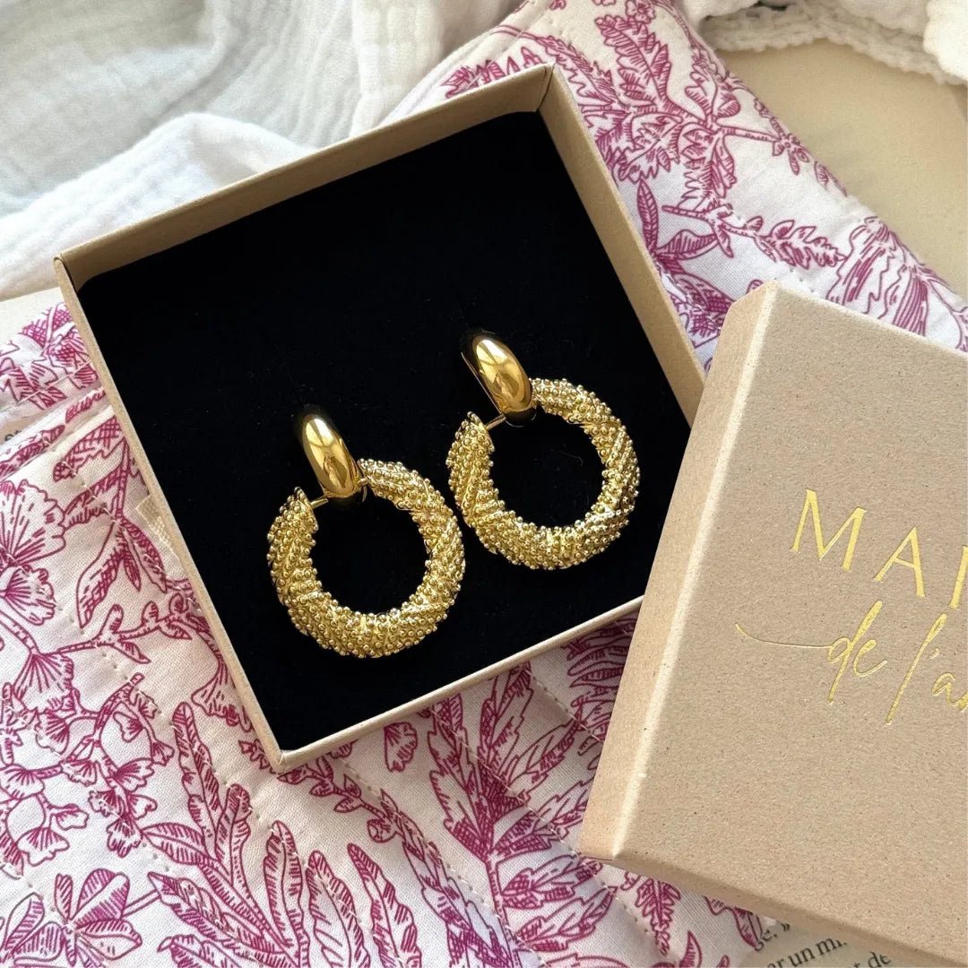 Paire de boucles d'oreilles créoles dorées granulées dans une boîte cadeau ouverte sur tissu blanc à motifs feuilles violets.