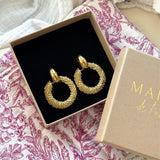 Paire de boucles d'oreilles créoles dorées granulées dans une boîte cadeau ouverte sur tissu blanc à motifs feuilles violets.