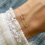 Gros plan bracelet fin en acier inoxydable doré avec motif double maillons rectangulaires entrelacés, dont un pavé de strass brillants.