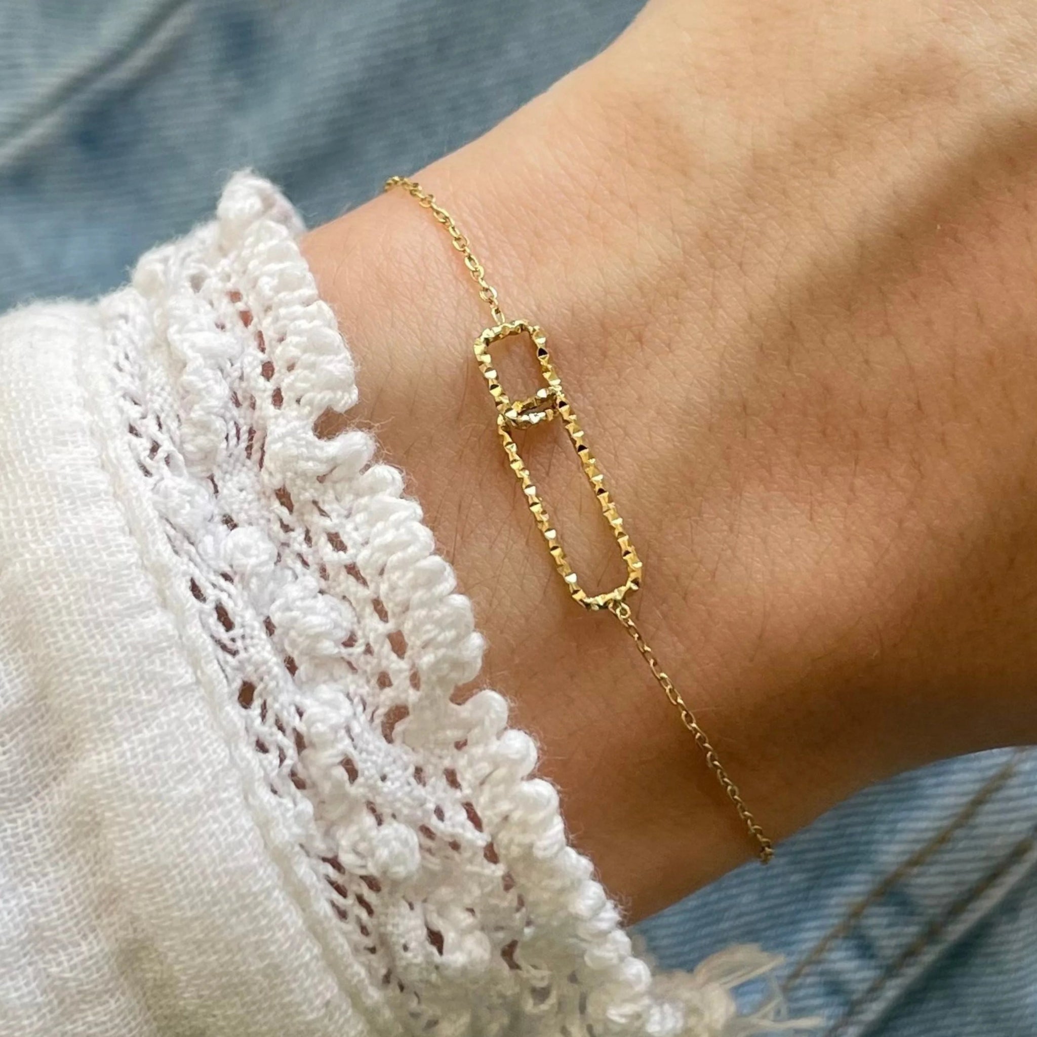 Gros plan bracelet fin en acier inoxydable doré avec motif double maillons rectangulaires entrelacés, dont un pavé de strass brillants.