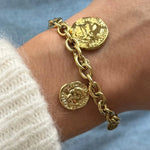 Gros plan bracelet grosse maille en acier inoxydable doré avec breloques pièces de monnaie antiques porté au poignet.
