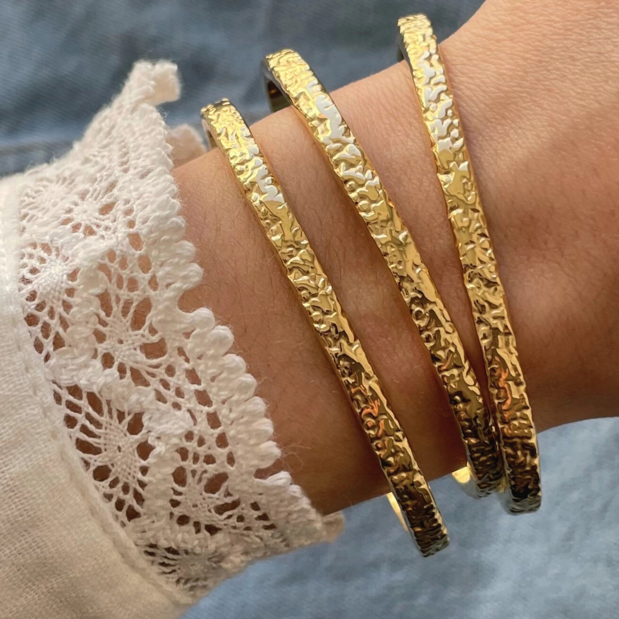 Accumulation de trois bracelets joncs ouverts rigides Azalea en acier inoxydable doré à effet martelé texturé.