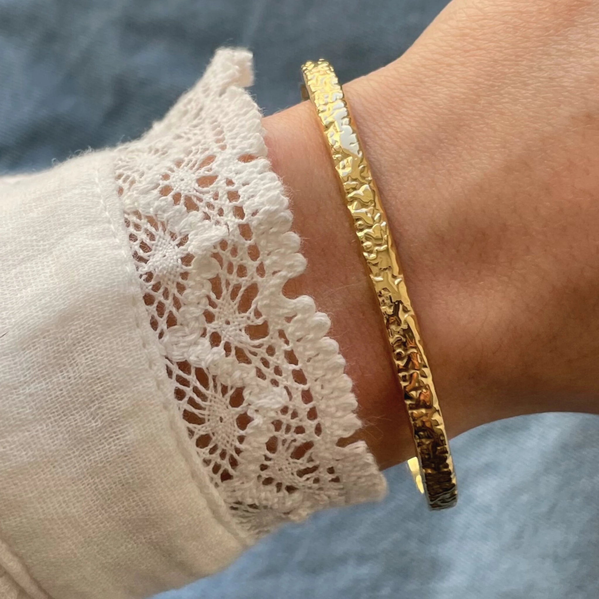 Bracelet jonc rigide fin et texturé doré porté seul au poignet sur une manche en dentelle.