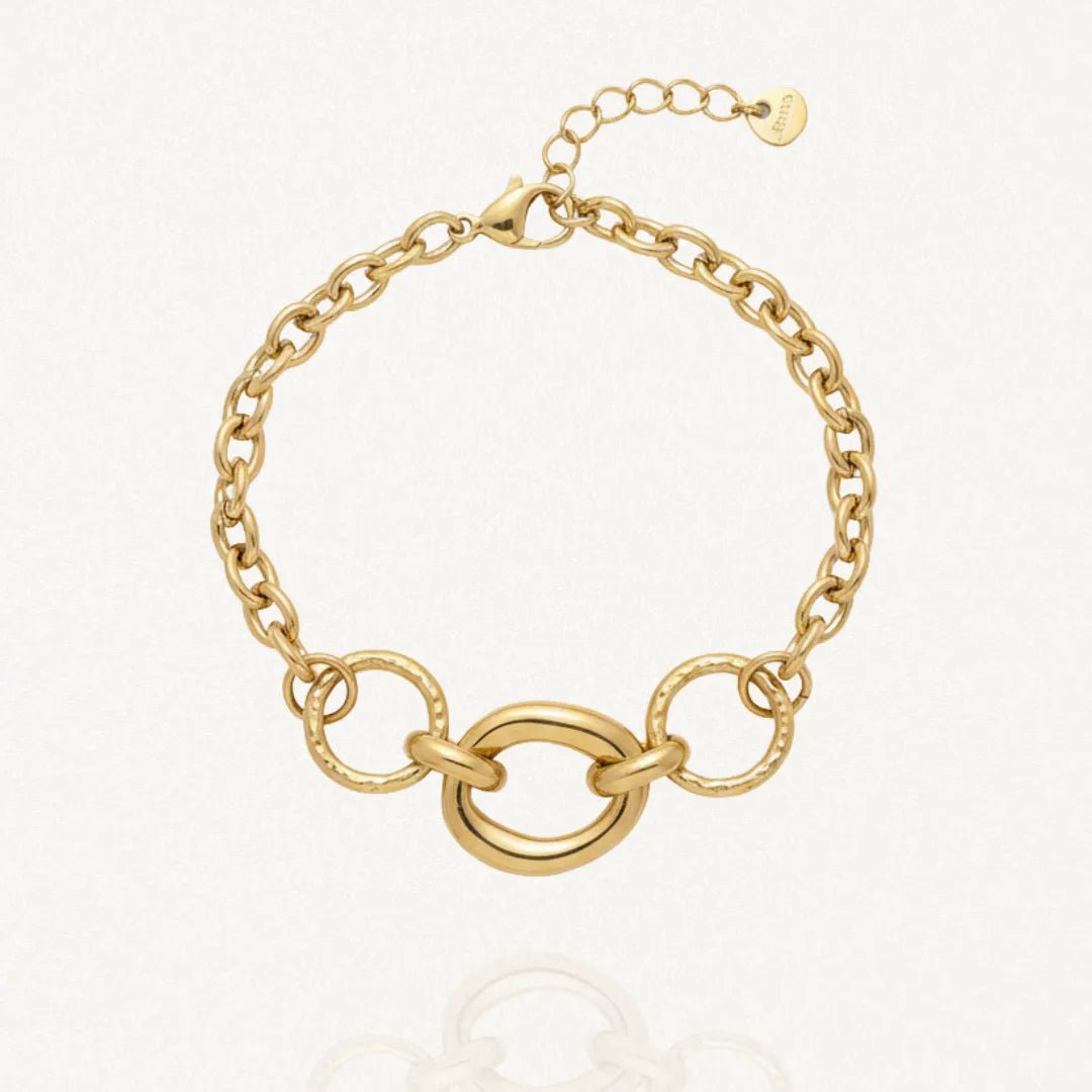 Bracelet en acier inoxydable doré composé de maillons épais et d'une section centrale avec un grand anneau ovale lisse encadré de deux anneaux ronds texturés. Fermoir mousqueton et chaînette d'extension.