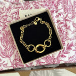 Bracelet doré, maillons ronds texturés et ovale lisse, fermoir mousqueton, breloque cœur, boîte noire sur tissu fleuri rose.