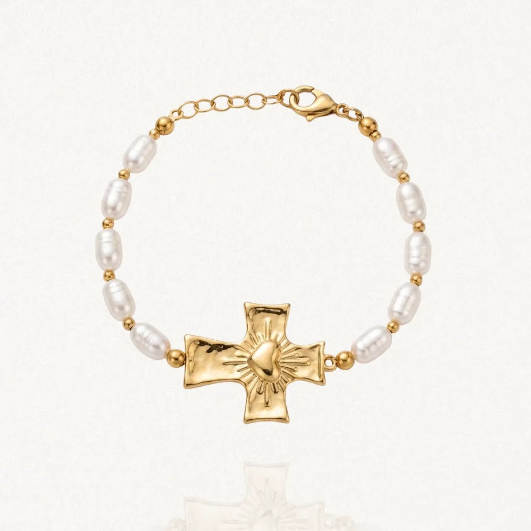 Bracelet doré composé de perles baroques blanches et de petites billes, centré sur une croix texturée avec un cœur rayonnant. Fermoir mousqueton et chaînette d'extension visibles.